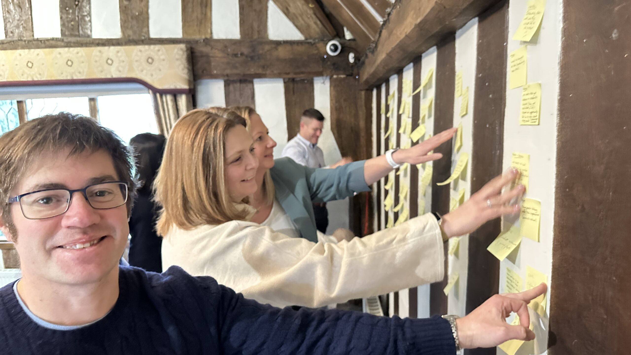 Participants engage in a workshop, placing yellow sticky notes on a wall in a room with wooden beams and warm lighting.