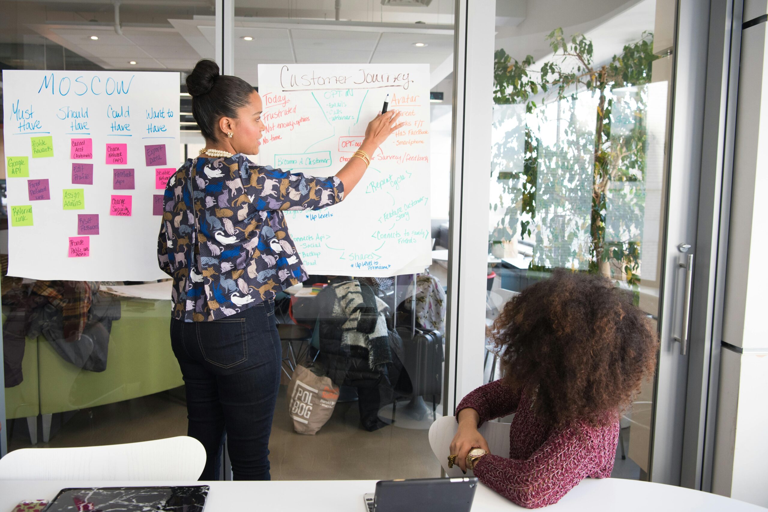 A woman in a patterned blouse writes on a whiteboard, while another woman with curly hair watches from a table with a laptop.