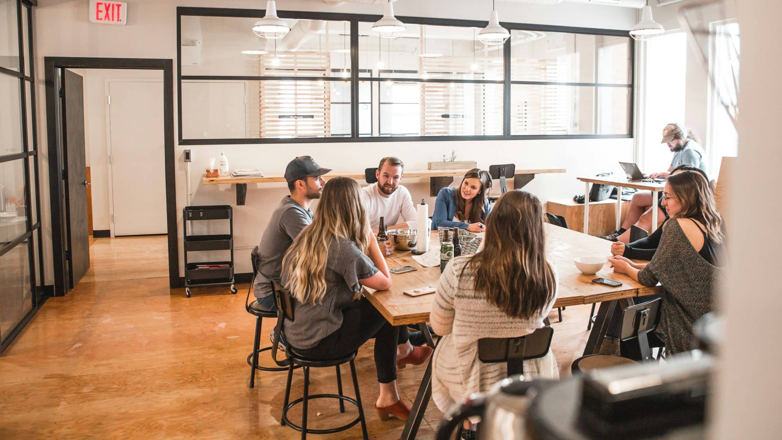 A group of eight people sits around a wooden table in a bright, modern room, engaging in conversation with drinks and notebooks.