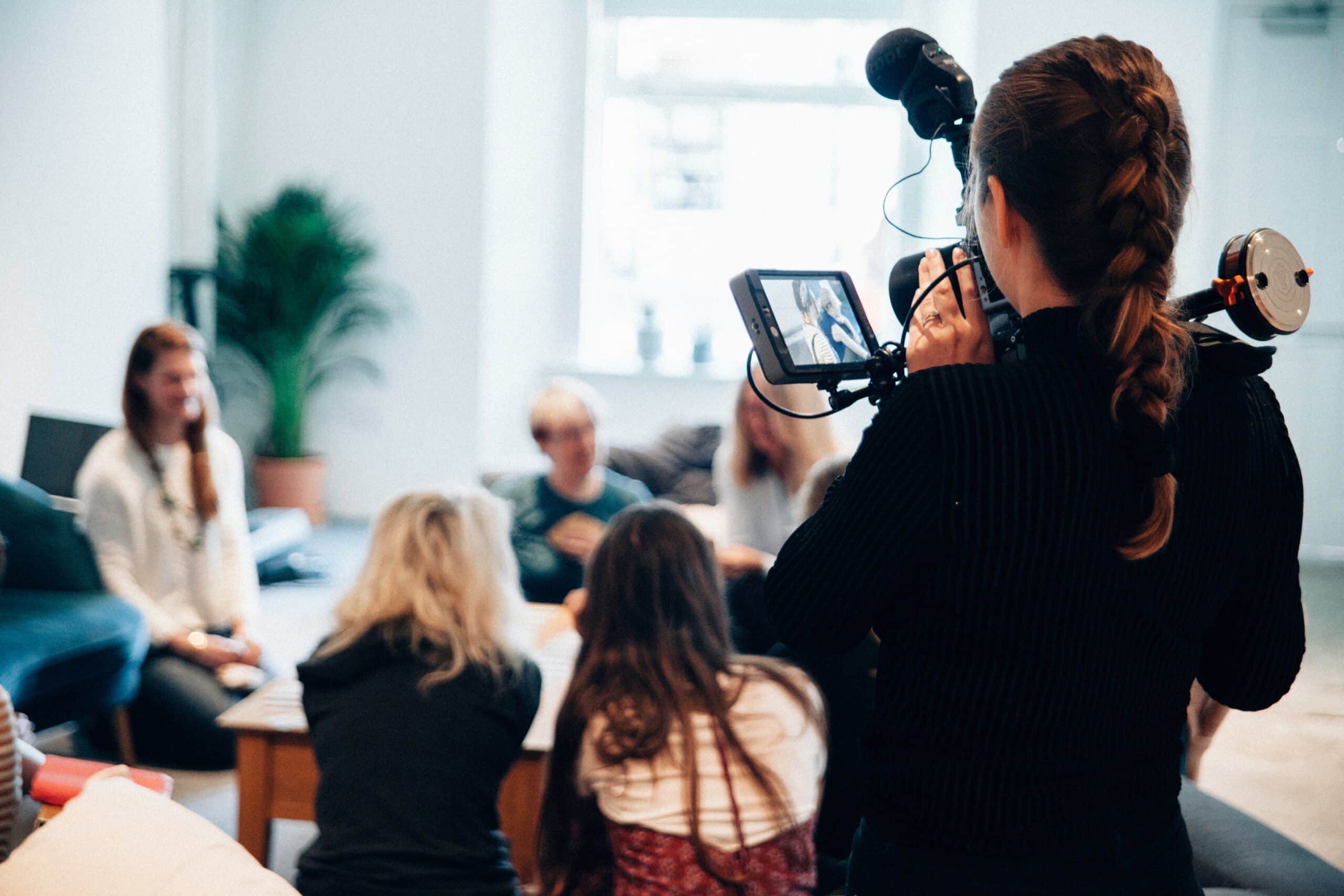 A person with a braided hairstyle holds a camera, filming a group of seated individuals engaged in discussion at a light-filled room.