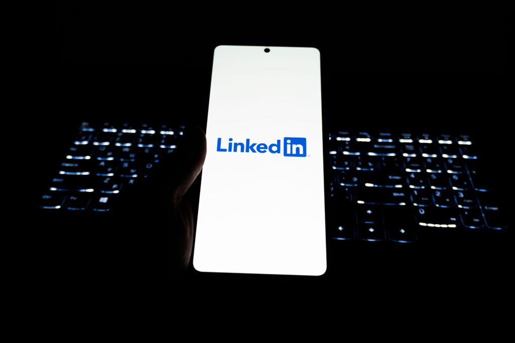 How LinkedIn and media activity intersect - Hand holding a smartphone displaying the LinkedIn logo, illuminated against a dark background with a softly glowing keyboard.