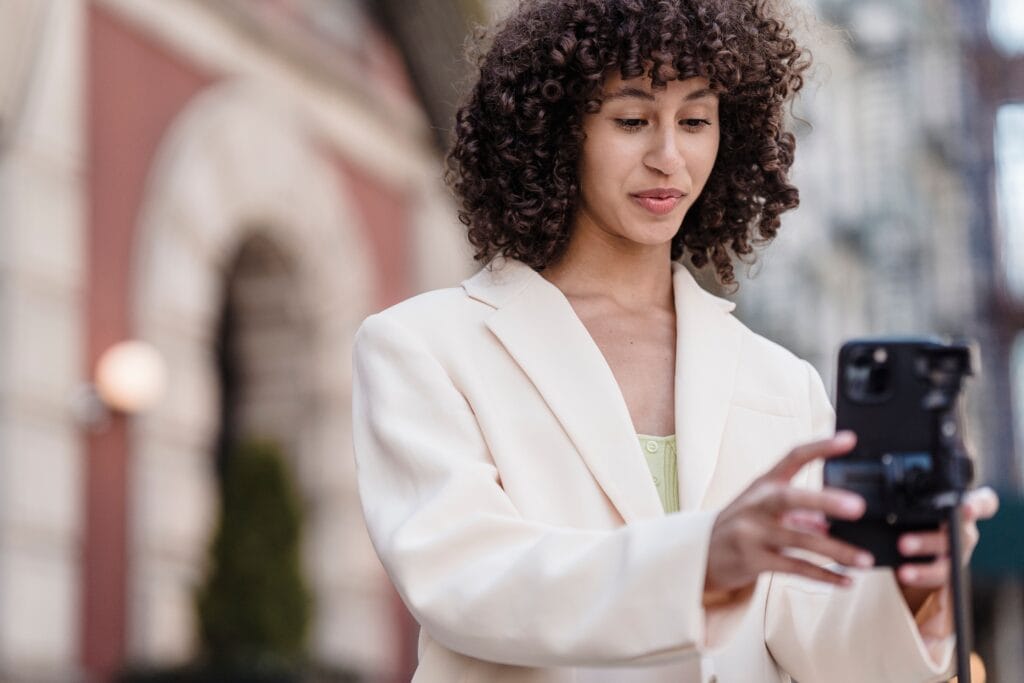 Avoid these common mistakes when creating in-house videos. Young woman with curly hair wearing a cream blazer, smiling while holding a smartphone outdoors in an urban setting.