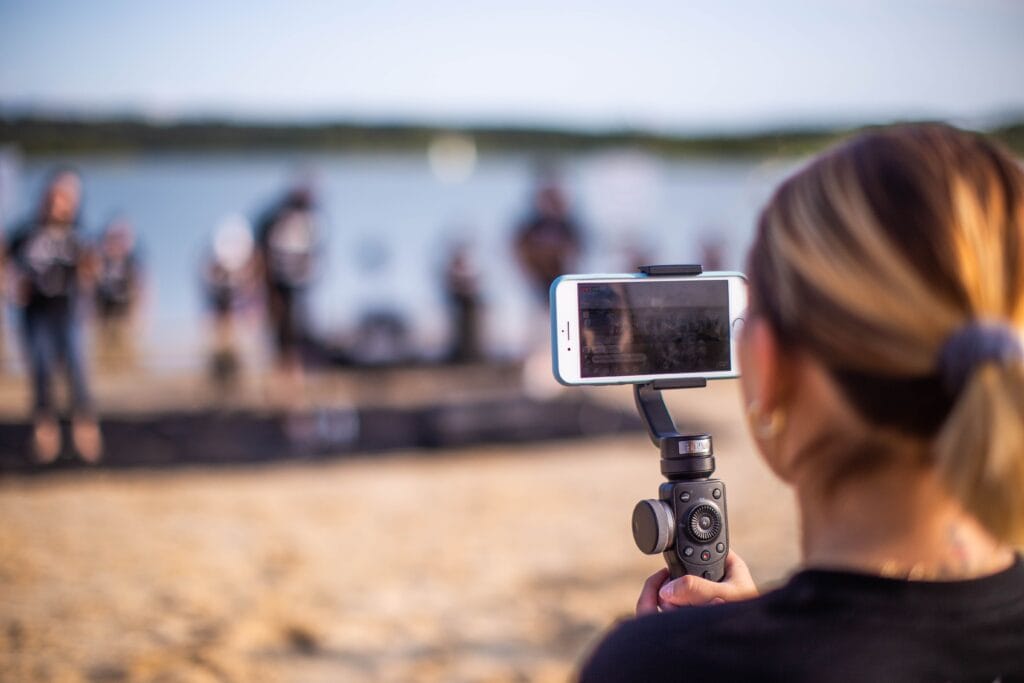 How non-video teams can create professional videos quickly. A person holds a smartphone on a stabiliser while filming a group performing on a beach, with water and trees in the background.