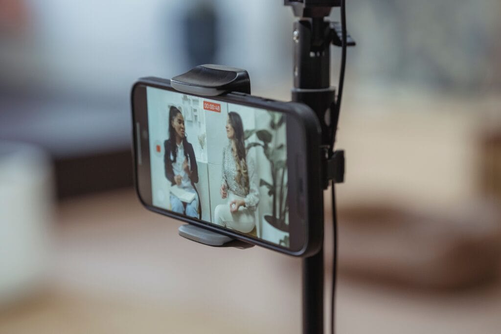 Why smartphone video is essential for communication. Smartphone mounted on a tripod records a conversation between two seated people in an indoor setting. The camera focuses on the phone screen, which shows the two speakers, while the surrounding room appears softly blurred in the background. A recording timer is visible on the phone display.