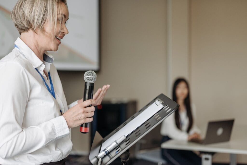 How education leaders can strengthen trust through the media - A woman speaking into a microphone stands beside a laptop, while another person seated at a desk listens attentively in a conference setting.
